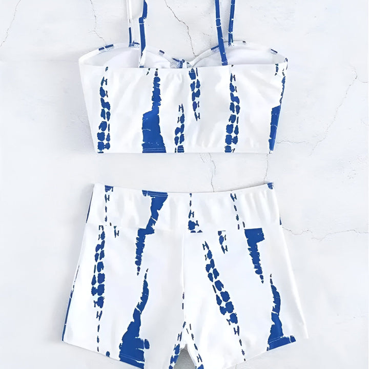 Une jeune femme blonde pose en maillot de bain deux pièces à motifs marron et blanc, devant une piscine turquoise, avec des arbres et un bâtiment blanc en arrière-plan, sous un ciel bleu ensoleillé.