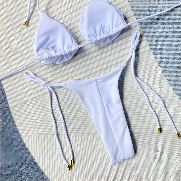 Une femme posant devant un mur de pierre porte un bikini blanc avec des détails floraux sur le haut. Le bas du bikini est noué sur les côtés.