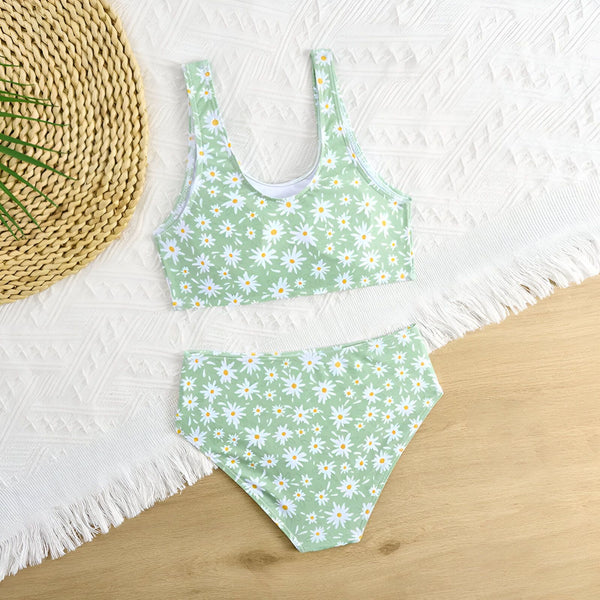 Culotte de maillot de bain vert clair à motif floral marguerite blanche et jaune, sur fond en bois clair. Une décoration en forme de coquillage est visible en bas à droite.