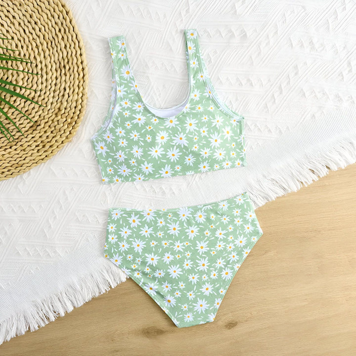 Culotte de maillot de bain vert clair à motif floral marguerite blanche et jaune, sur fond en bois clair. Une décoration en forme de coquillage est visible en bas à droite.