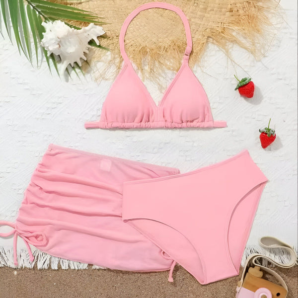 Vue rapprochée d'un ensemble de bikini rose avec un haut triangle, une culotte taille haute et une jupe portefeuille assortie, accompagné d'un chapeau de paille sur un fond de plage. Parfait pour l'été et les vacances.