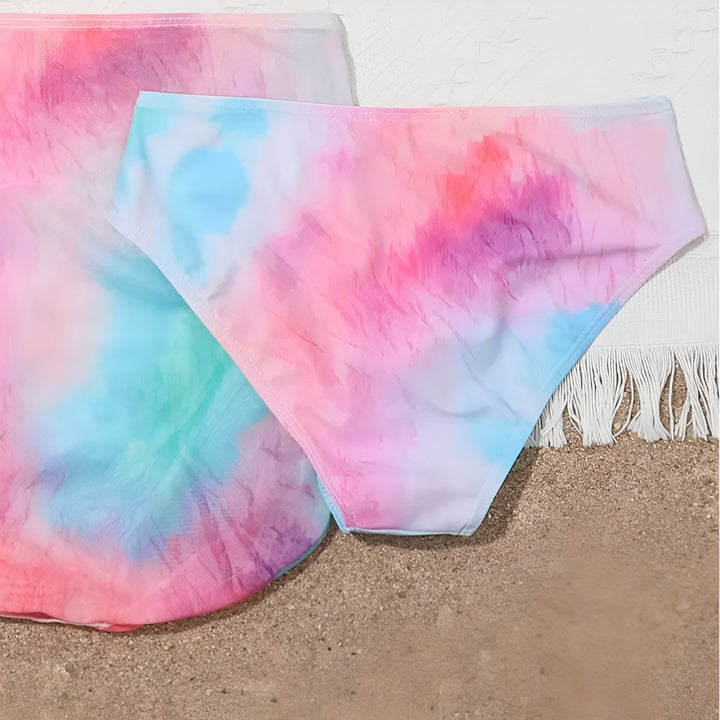 Ensemble de plage tie-dye multicolore sur une serviette à franges, posé sur du sable. Vêtements d'été colorés, tendance boho chic.
