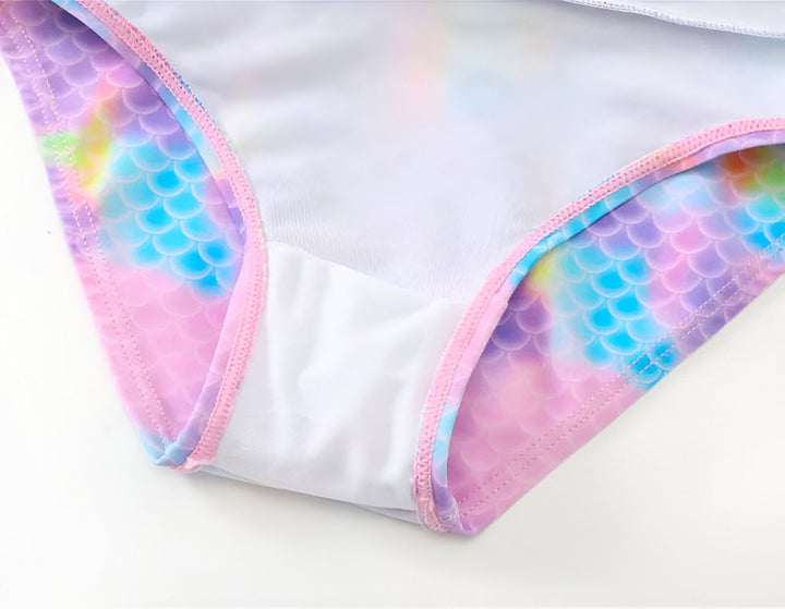 Maillot de bain une pièce pour enfant avec flotteurs intégrés et bonnet assorti, motif écailles de sirène multicolores, coquillages et palme décorative sur fond blanc. Vêtement de plage pour enfant.