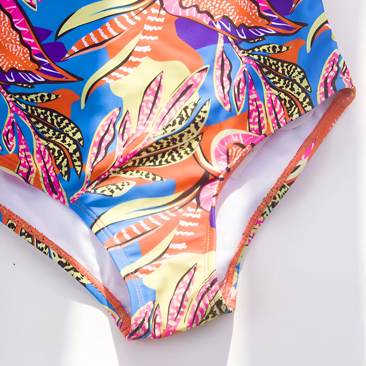 Gros plan d'un bas de maillot de bain à motifs tropicaux colorés (bleu, orange, jaune, rose) sur fond blanc. Détails de la bordure orange. Idéal pour la mode estivale, la plage et les vacances.