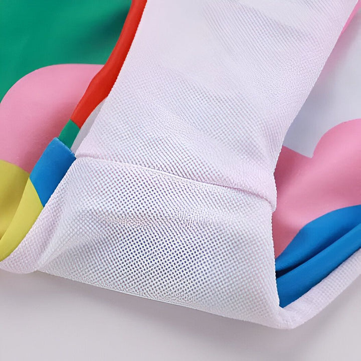 Maillot de bain deux pièces pour enfant, motif nuages colorés (rouge, bleu, rose, vert, jaune) avec un volant asymétrique sur le haut. Chapeau de soleil et décorations autour. Mode enfantine, été.