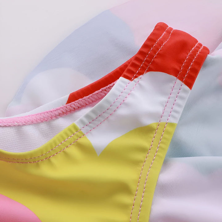 Gros plan d'un tissu coloré avec des motifs en forme de nuage. Les couleurs incluent le rose, le rouge, le bleu, le vert, le jaune et le blanc. Le tissu a un bord à volants.