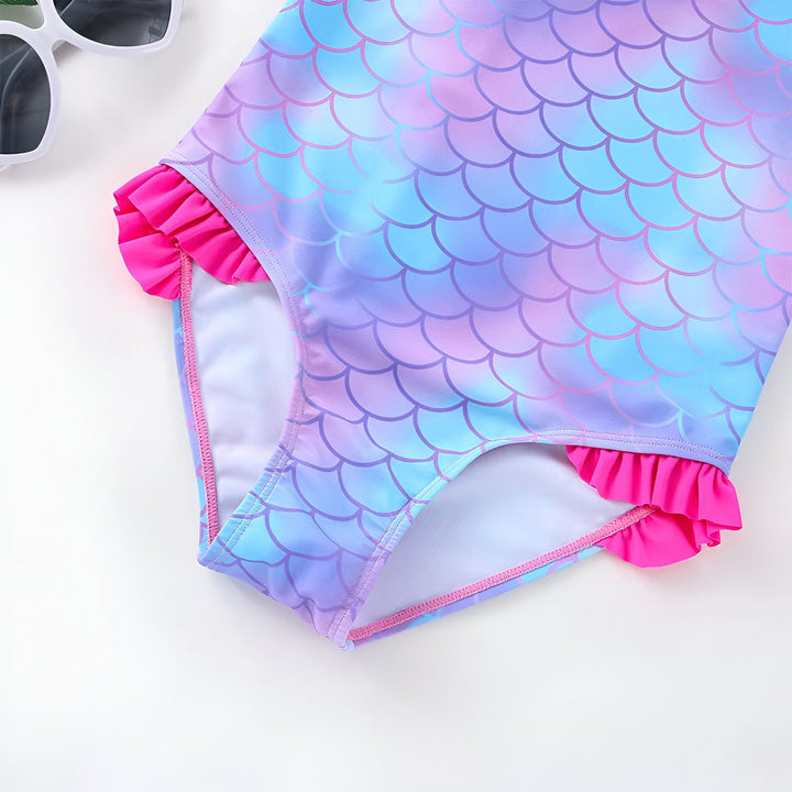 Gros plan d'un maillot de bain une pièce pour enfant avec motif écailles de sirène bleu et violet, bordé de volants rose vif. Des lunettes de soleil blanches sont partiellement visibles sur le côté.