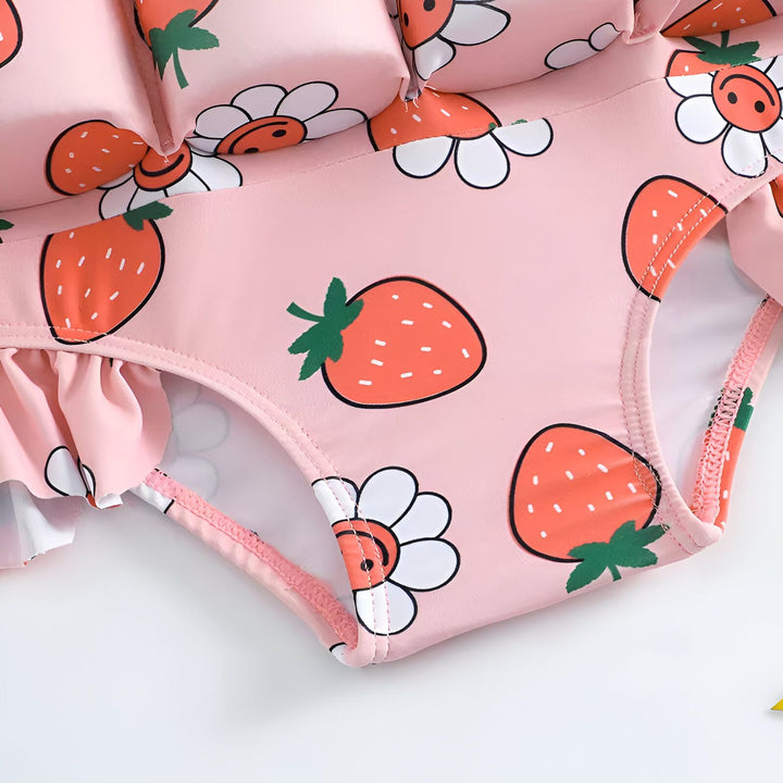 Gros plan sur un maillot de bain rose pour enfant, orné de motifs de fraises et de marguerites souriantes. Des volants roses ajoutent une touche fantaisiste.