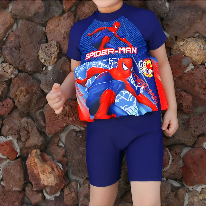 Un jeune garçon en maillot de bain bleu Spiderman avec une aide à la flottabilité devant un mur de pierres. Prêt pour la piscine ou la plage. Vêtements de bain pour enfants, été.
