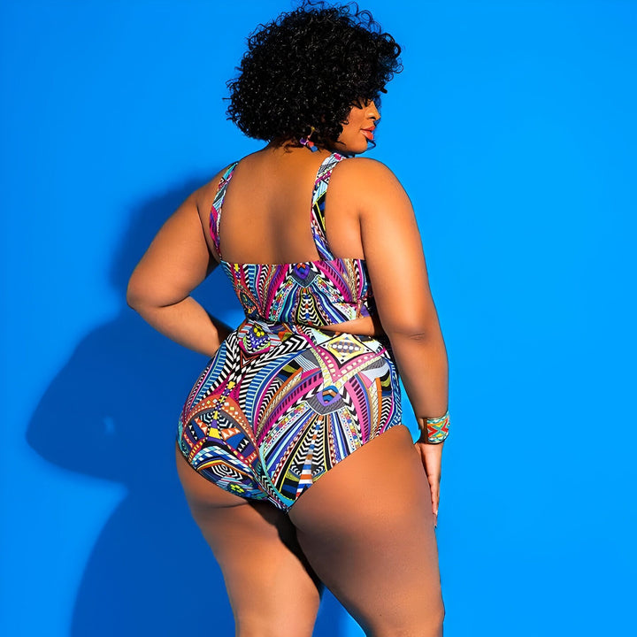 Une femme afro-américaine vue de dos, portant un maillot de bain deux pièces à motifs géométriques multicolores sur fond bleu uni. Elle a des cheveux bouclés noirs et des boucles d'oreilles colorées. Un bracelet est visible à son poignet. Mode estivale.