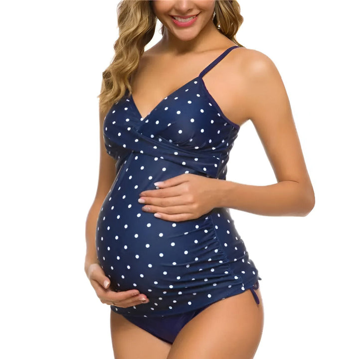 Maillot de bain de grossesse noir 2 pièces avec haut tankini croisé et culotte assortie. Bretelles fines et détails à nouer sur les côtés. Femme souriante avec une main dans les cheveux.