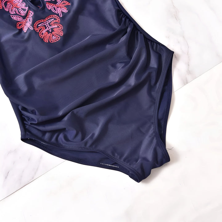 Femme enceinte portant un maillot de bain une pièce bleu marine avec des broderies de feuilles roses et rouges sur le devant, col en V avec lacets, fronces sur les côtés. Idéal pour la maternité à la plage ou à la piscine.