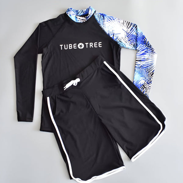 Un jeune homme souriant porte un rashguard noir à manches longues avec motif de feuilles de palmier sur une manche, des lunettes de soleil et un short de bain noir sur une plage tropicale ensoleillée.