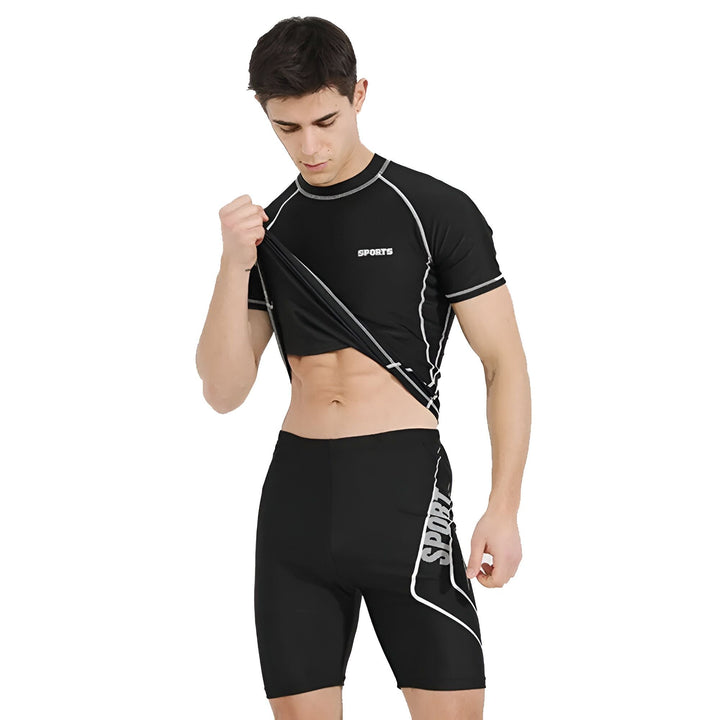 Homme en tenue de sport noire, t-shirt moulant "SPORTS" et short "SPORT", étirant le tissu du t-shirt pour montrer son élasticité. Idéal pour l'entraînement, la natation ou la course. Style athlétique et confort.