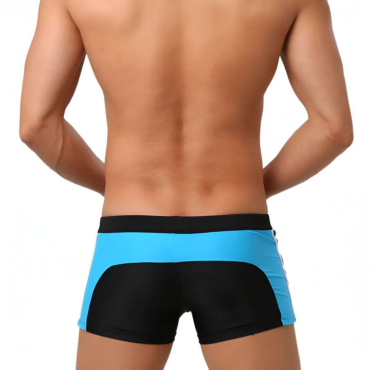 Homme torse nu portant un short de bain noir et bleu avec des cordons blancs. Détail d'une fermeture éclair blanche sur le short. Silhouette athlétique visible.