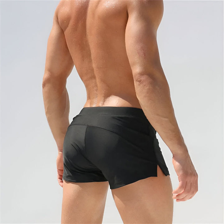 Homme torse nu en short de bain noir avec cordon de serrage, main dans la poche. Idéal pour la mode masculine, les vacances et le style de vie estival.