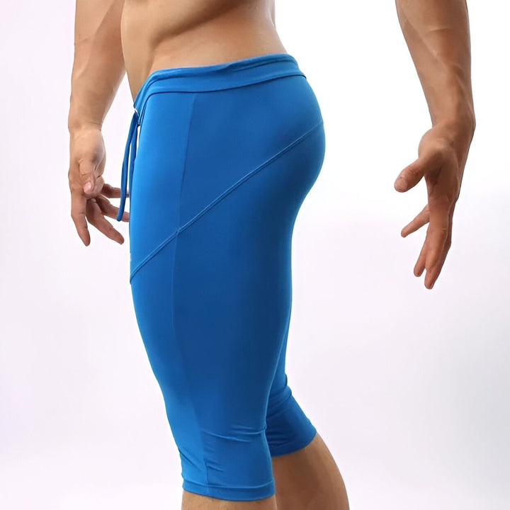 Homme en tenue de sport, portant un corsaire vert olive moulant. Le pantalon arrive en dessous des genoux et a un cordon de serrage à la taille. Les mains sur les hanches, les abdos apparents. Vêtement de sport pour homme.