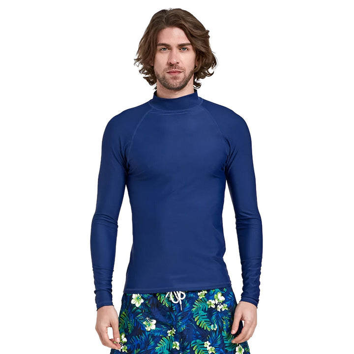 Homme portant un rashguard bleu à manches longues et un short de bain floral, debout devant un fond blanc. Vêtements de protection solaire pour la plage.