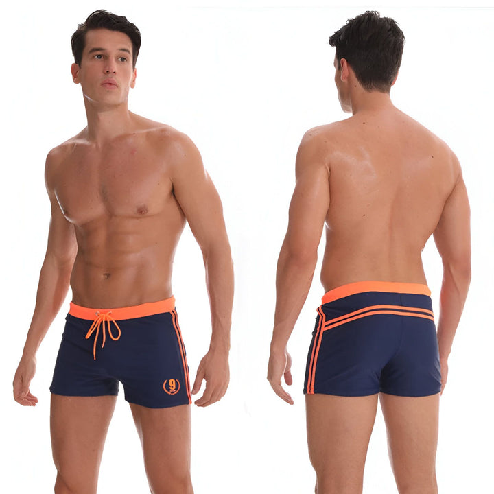 Homme torse nu portant un slip de bain bleu marine avec taille et rayures orange. Modèle athlétique, regardant sur le côté.