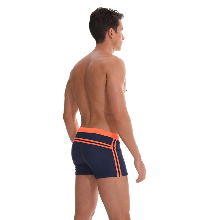 Homme torse nu portant un slip de bain bleu marine avec bordure et rayures orange, et le logo "9" sur la jambe.