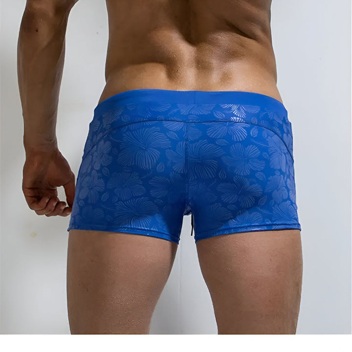 Un homme portant un short de bain bleu à motifs floraux, avec une main dans la poche.