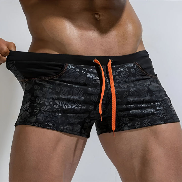 Image rapprochée d'un homme portant un slip de bain bleu à motifs floraux, main dans la poche. Détail des abdominaux et des bras musclés. Idéal pour la mode masculine et la collection été.