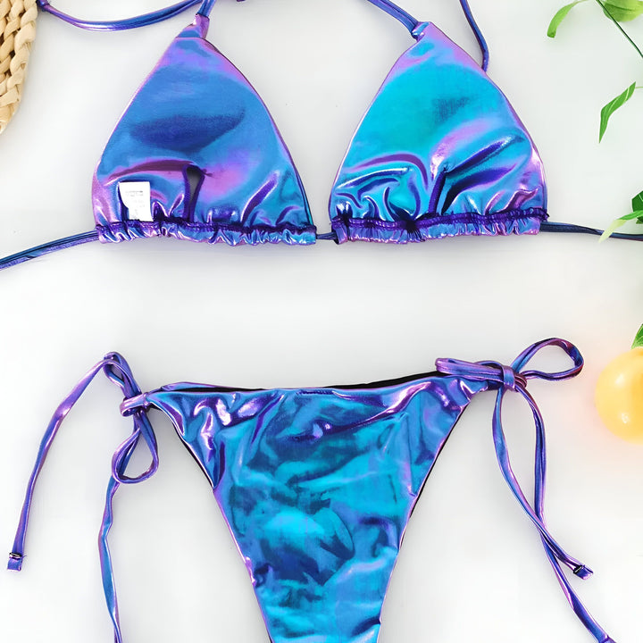 Image d'un bikini deux pièces bleu iridescent, haut triangle à nouer et bas échancré à nouer sur fond blanc. Accessoires décoratifs : chapeau de paille, balle jaune et feuille verte. Idéal pour la plage et la piscine.