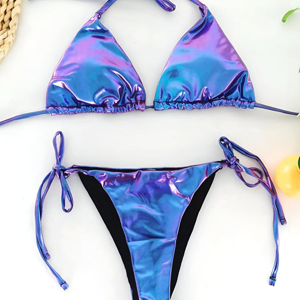 Image d'un bikini deux pièces irisé bleu et violet, présenté avec des accessoires de mode et des éléments de nature morte sur un fond blanc. Idéal pour le style d'été et la plage.