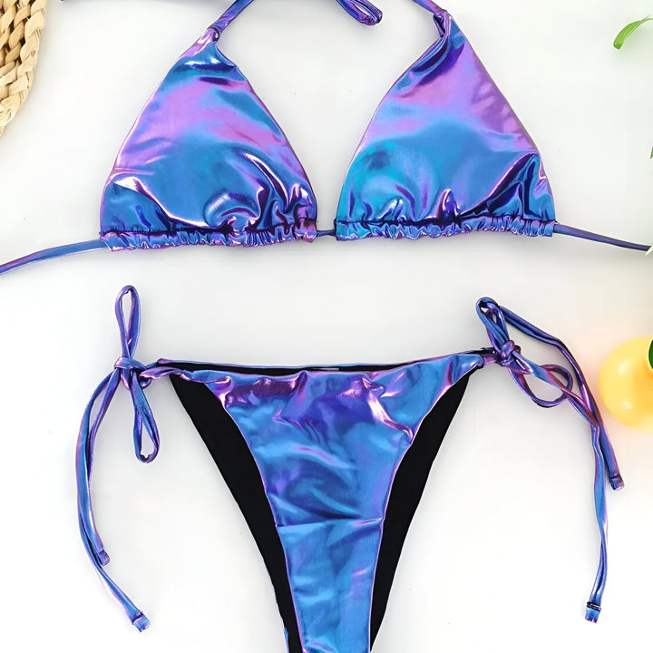 Image d'un bikini deux pièces bleu iridescent, comprenant un haut triangle à lacets et une culotte assortie, sur un fond blanc avec une décoration naturelle. Maillot de bain tendance.