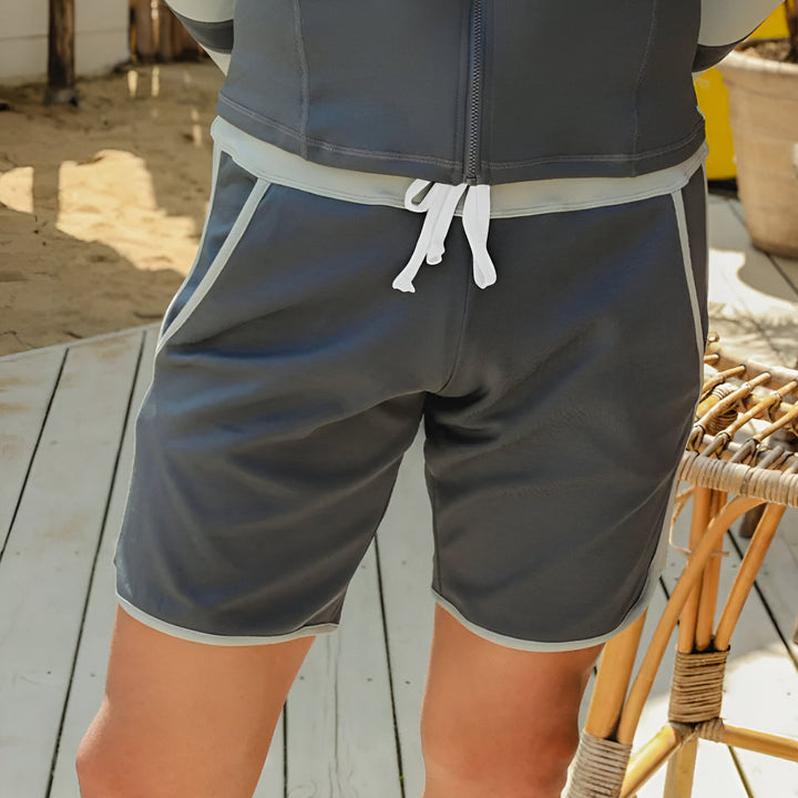 Gros plan d'un short de bain gris avec cordon blanc et bordure claire. On voit une partie du haut de bain assorti et une chaise en osier en arrière-plan, suggérant une scène de plage ou de piscine.