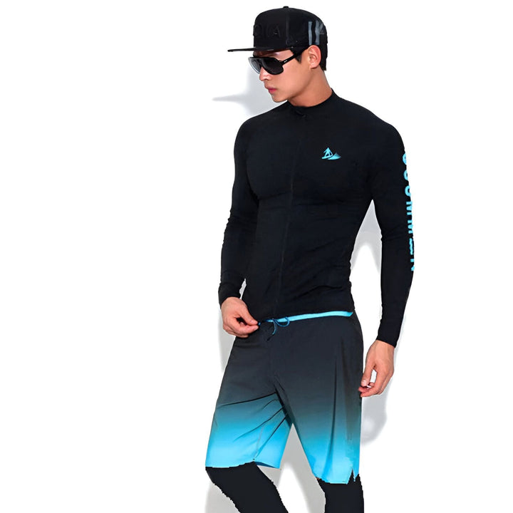 Homme portant un rashguard noir avec un logo turquoise, un short de bain noir et une casquette noire. Il porte des lunettes de soleil et pose devant un fond blanc.