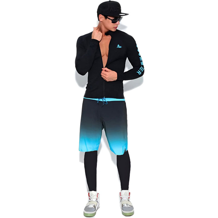 Un homme avec une casquette et des lunettes de soleil tient une planche de surf blanche. Il porte un haut noir à manches longues, un short dégradé bleu et noir, des leggings noirs et des baskets.