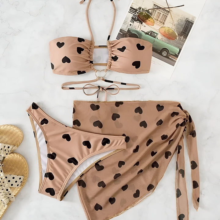 Femme portant un bikini et un paréo beiges avec des motifs de cœurs noirs, tenue d'été à la mode, prêt pour la plage.