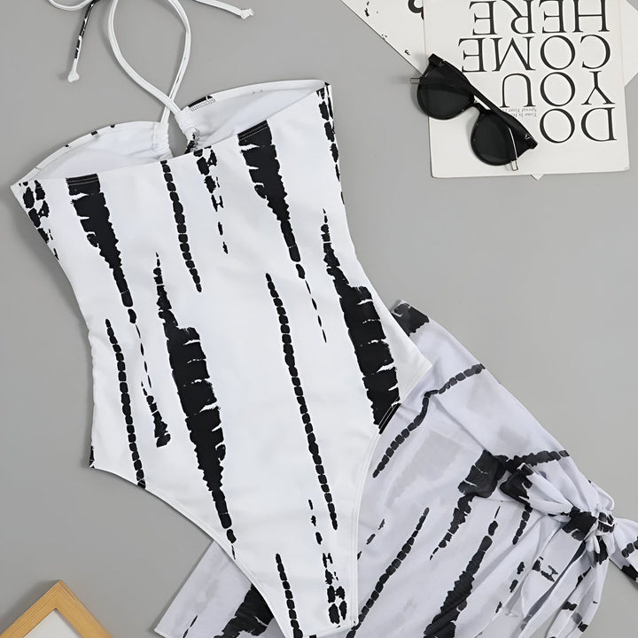 Une femme porte un maillot de bain une pièce noir et blanc à imprimé abstrait, avec un décolleté plongeant et une jupe nouée sur le côté, mettant en valeur sa silhouette élégante. Idéal pour la plage ou la piscine.