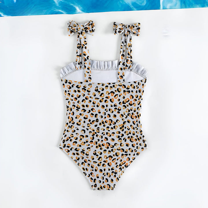 Maillot de bain une pièce pour fille avec imprimé léopard, bretelles à nœuds, volant sur le haut. Sur un fond blanc avec une feuille de palmier, des tranches de pastèque et un coquillage.