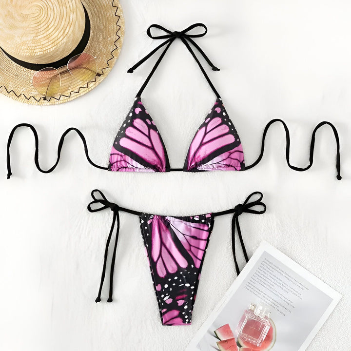 Femme souriante en bikini à motif papillon sur une plage de sable, portant une bague et un bracelet dorés. Mode estivale, vacances à la plage.