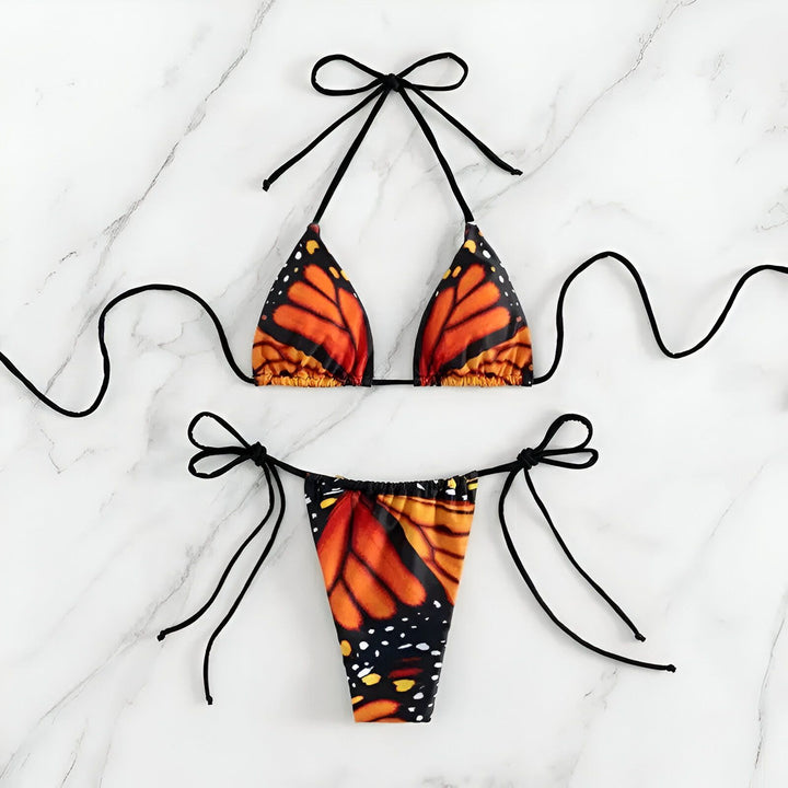 Une femme en bikini motif papillon monarque sur une plage ensoleillée, arborant un collier ras du cou noir. Le fond flou révèle des parasols et des chaises longues, créant une ambiance estivale.