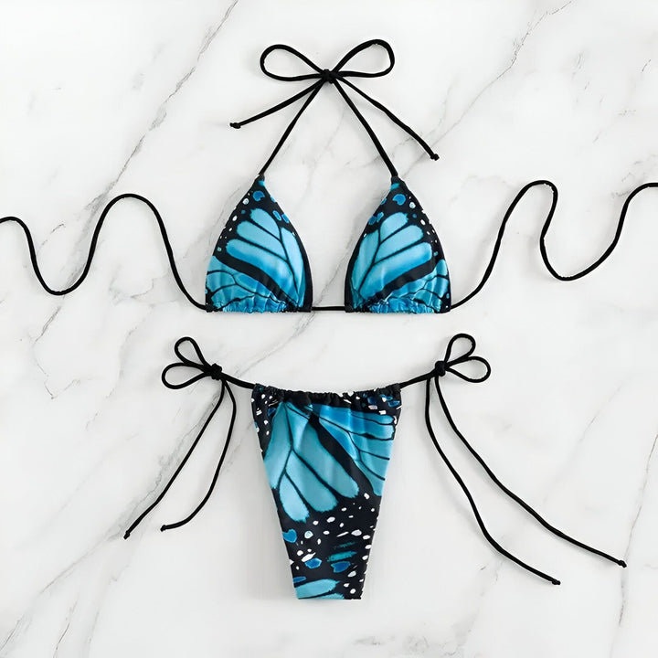 Vue arrière d'une femme sur la plage, portant un bikini string avec un motif de papillon bleu, arborant des tatouages d'un arbre avec une silhouette, de l'Œil d'Horus et d'une rose.