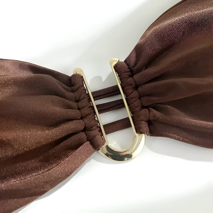 Image d'une femme portant un bikini marron sans bretelles avec des bijoux dorés sur une plage de sable blanc. Accessoires de mode estivale et bronzage parfait.
