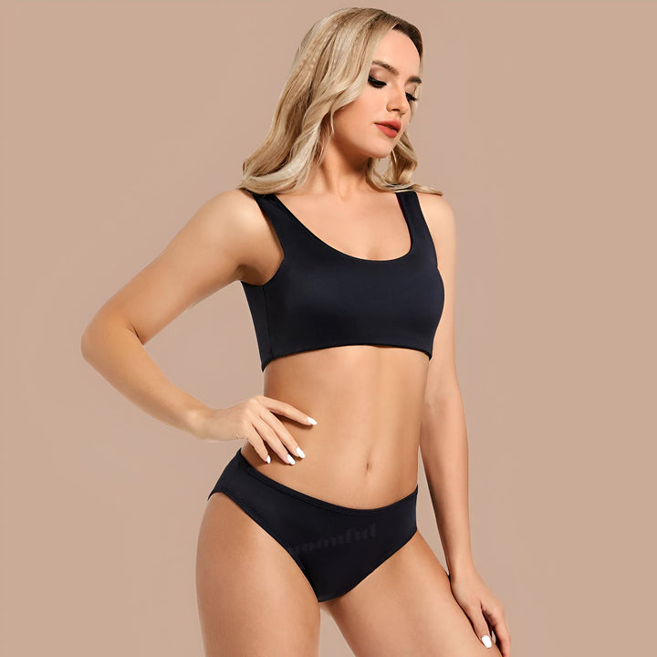Femme blonde en ensemble de sous-vêtements noir, pose sur fond beige. Soutien-gorge de sport et culotte assortie. Cheveux ondulés, regard vers le bas. Ongles blancs.