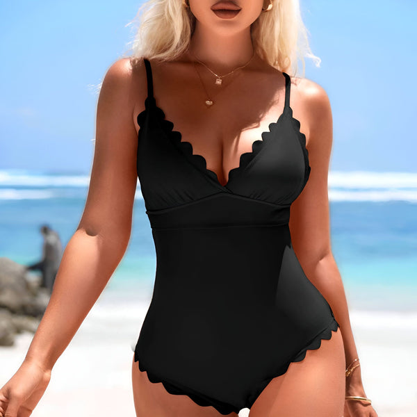 Une femme blonde, portant un maillot de bain noir à volants, un chapeau de paille et des bijoux en or, pose avec un sac de plage en osier devant un ciel bleu et l'océan turquoise.