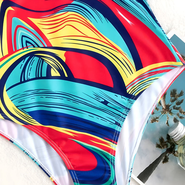 Femme en maillot de bain une pièce coloré avec motif abstrait et détails bijoux, posant avec une veste en jean claire sur un lit blanc. Accessoires modernes et ambiance décontractée.