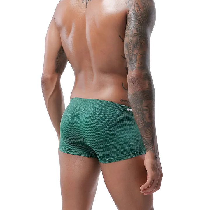 Homme torse nu, athlétique, portant un maillot de bain bleu marine à pois blancs, avec un cordon blanc à la taille et un tatouage sur le bras. Mode masculine estivale.