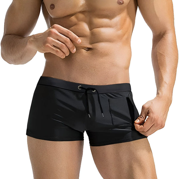 Homme musclé torse nu, portant un maillot de bain boxer noir avec cordon de serrage et poche latérale. Abdominaux dessinés, peau hâlée. Mode masculine, vêtement de plage.