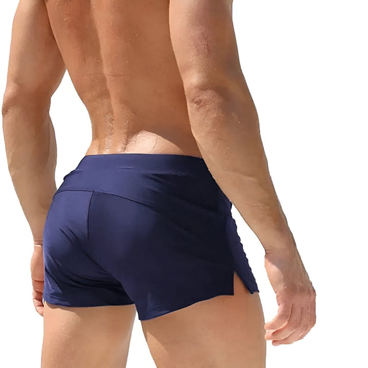 Gros plan de deux maillots de bain pour homme : un noir et un bleu violet, avec des cordons noirs noués. Détail des coutures et des œillets métalliques. Mode masculine estivale.