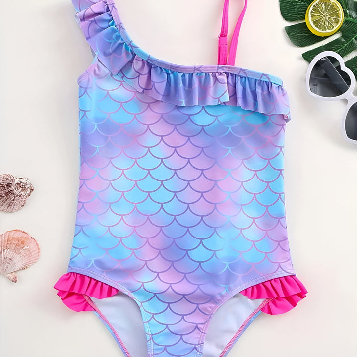 Gros plan sur un maillot de bain une pièce enfant, motif écailles de sirène bleu et violet, avec une bretelle rose et volant. Décoration tropicale : feuille de palmier et citron.