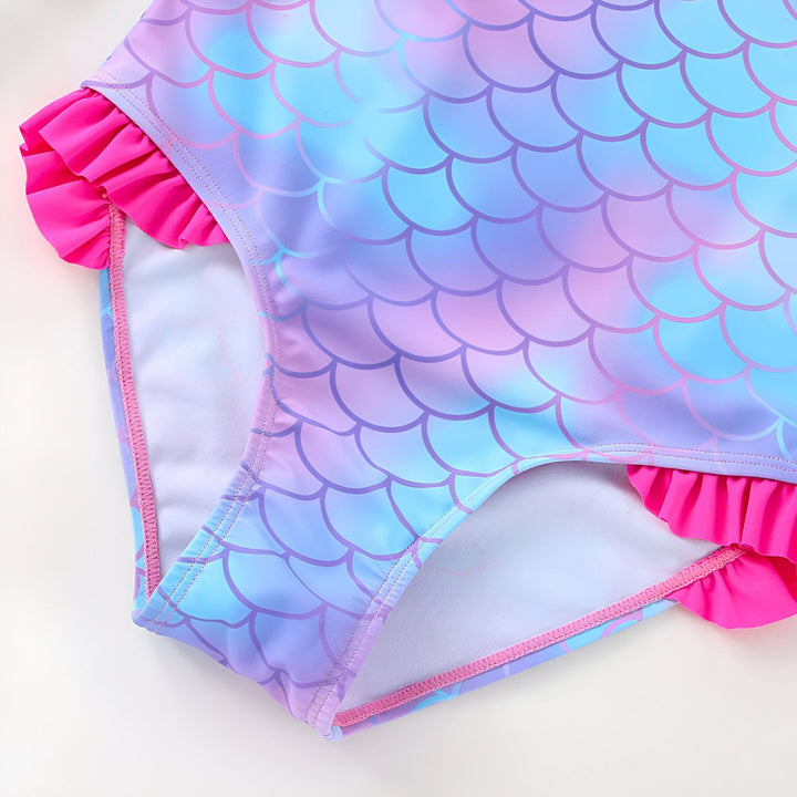 Gros plan d'un maillot de bain une pièce pour enfant avec un motif d'écailles de sirène bleues et violettes, agrémenté de volants roses vifs sur les côtés. Idéal pour les vacances d'été à la plage ou à la piscine.
