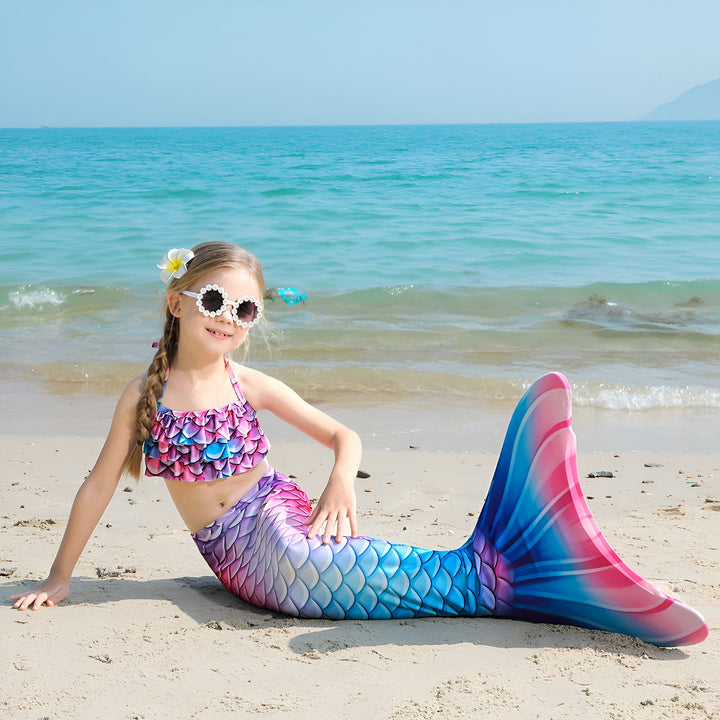 Une jeune fille souriante, déguisée en sirène avec une queue colorée rose et bleue, assise sur une plage de sable. Elle porte un haut de maillot de bain à écailles, des lunettes de soleil rondes et une fleur dans les cheveux.