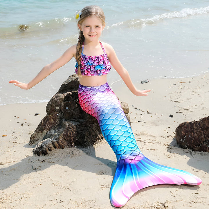 Fille en costume de sirène coloré sur une plage ensoleillée. Elle sourit et pose avec une queue de poisson aux nuances de rose, violet et bleu. Une fleur orne ses cheveux blonds tressés.