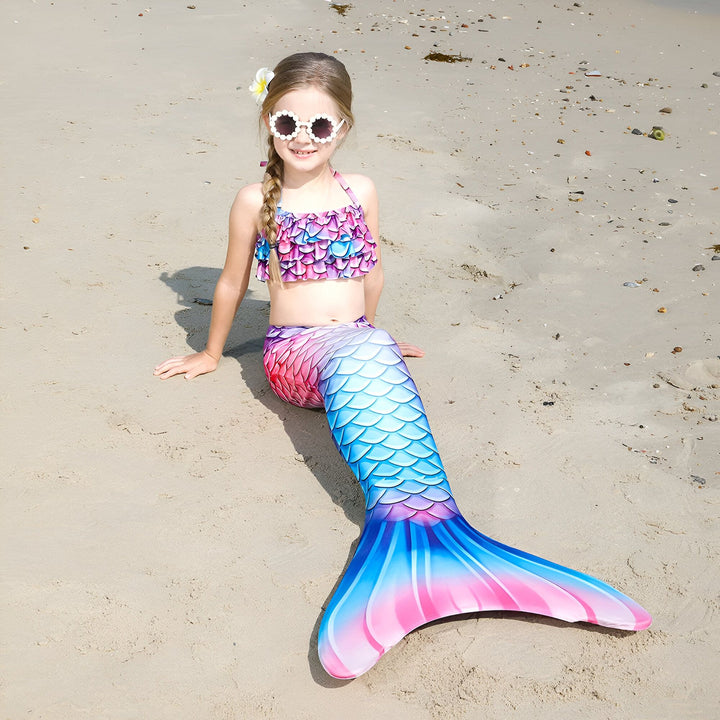 Une jeune fille souriante, en costume de sirène aux écailles colorées rose et bleu, est allongée sur une plage de sable fin avec l'océan turquoise en arrière-plan. Elle porte des lunettes de soleil fantaisie et une fleur dans les cheveux.